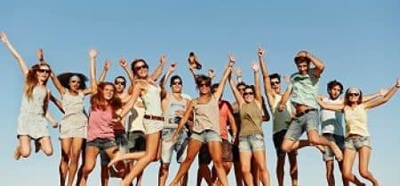 Group portrait of cheerful multi-ethnic friends jumping at beach. Full length of excited males and females in mid-air against clear blue sky. They are in casuals and sunglasses during summer vacation. (Group portrait of cheerful multi-ethnic friends j
